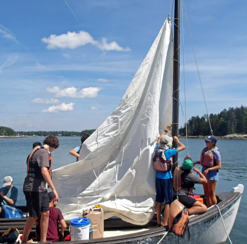 A group of people are gathered on a small sailboat, working together to raise the sail. The boat is on the water, with land visible in the background. The sky is partly cloudy. The people appear to be enjoying a sunny day out on the water, engaged in a sailing activity.